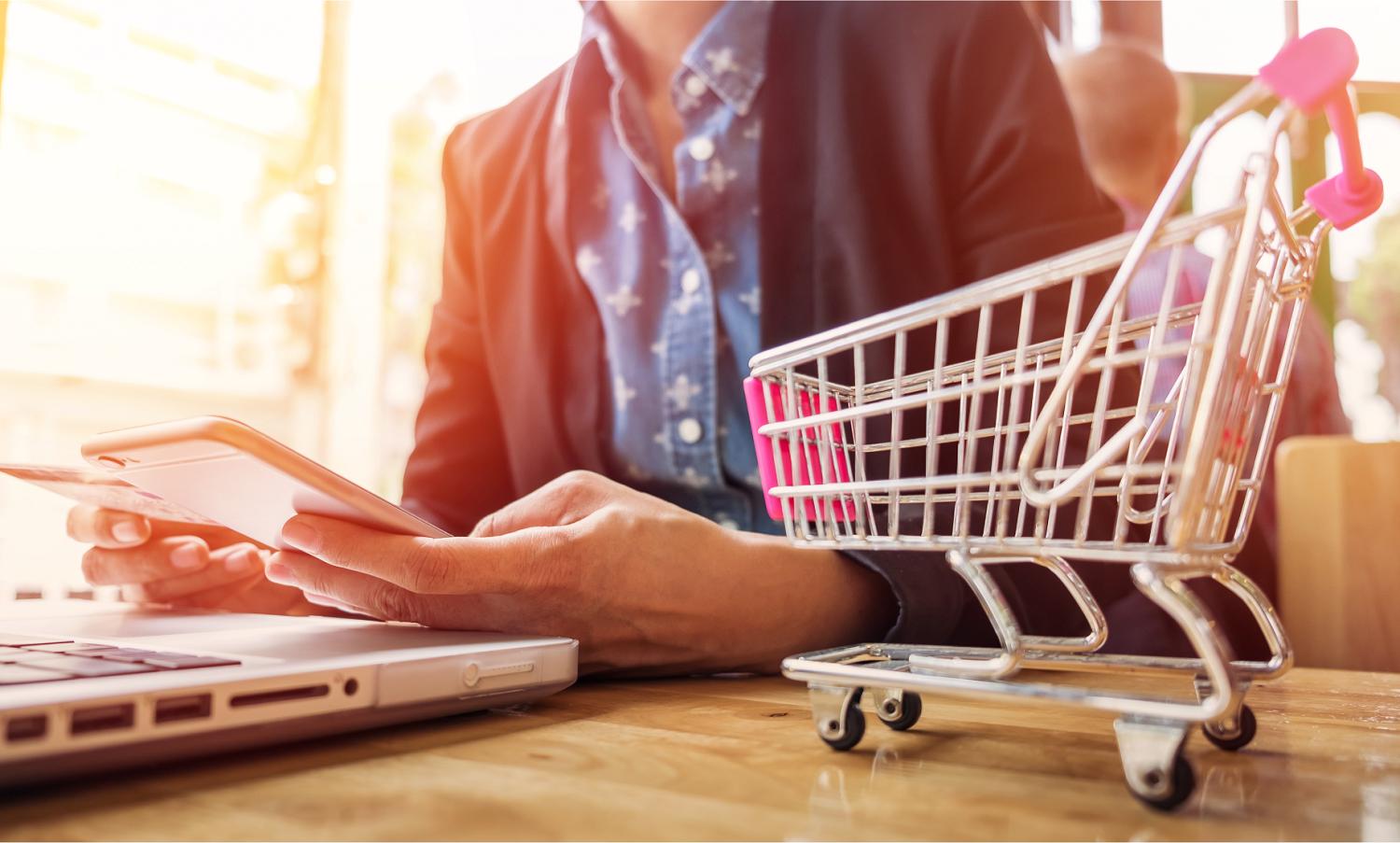 pessoa com um notebook na mesa, celular na mão e um carrinho de compras ao lado onde simula o futuro do ecommerce em 2023