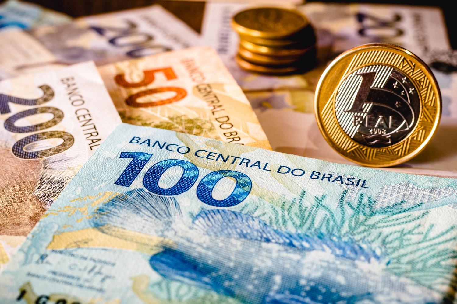 Notas de cem, duzentos e cinquenta reais e moedas brasileiras sobre uma mesa, simbolizando o pagamento de impostos e o impacto na sociedade.